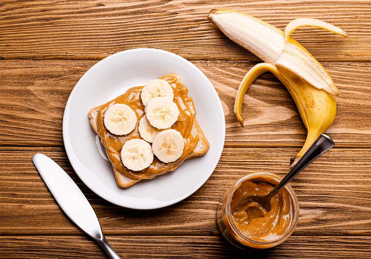 A slice of toast with peanut butter and banana slices on a white plate, next to a partially peeled banana and a jar of peanut butter—perfect for enjoying Retirement Living comfort at MorningStar Senior Living Kirkland, all set on a wooden table.