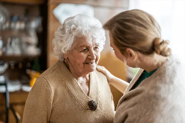 An elderly woman with white hair and a beige sweater smiles warmly as a younger woman gently holds her shoulder, both engaged in a caring conversation indoors at MorningStar Senior Living Kirkland, exemplifying compassionate retirement living.