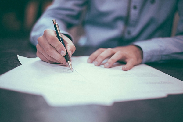 A person in a light blue shirt is writing on a stack of papers with a black and gold pen. The focus is on hands and documents, capturing the organized environment often found at MorningStar Senior Living Kirkland. The background is softly blurred.