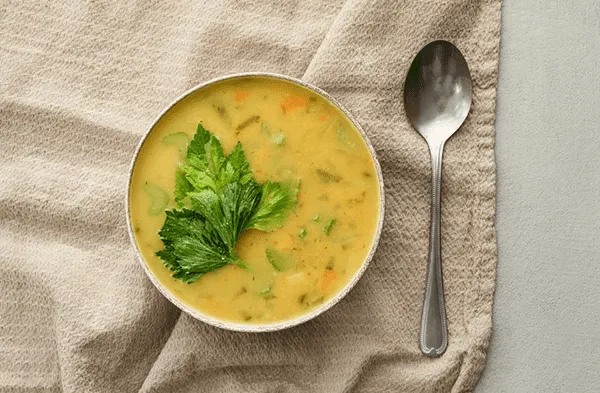 A bowl of creamy vegetable soup topped with fresh celery leaves sits on a beige cloth next to a silver spoon, offering a taste of comfort and warmth enjoyed at MorningStar Senior Living Kirkland—a place where retirement living feels like home.