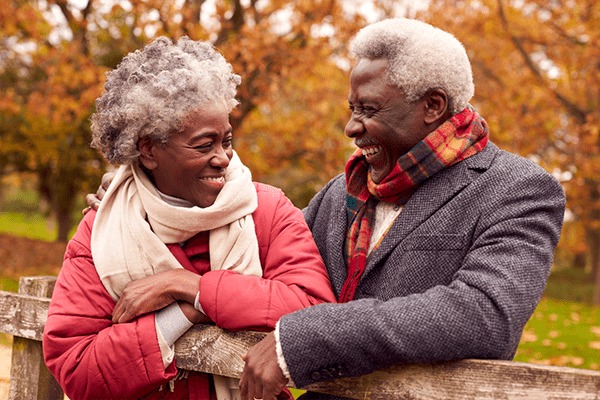 An older couple stands outdoors by a wooden fence, smiling warmly at each other. Enjoying Retirement Living, they wear autumn jackets and scarves, with colorful fall trees in the background.
