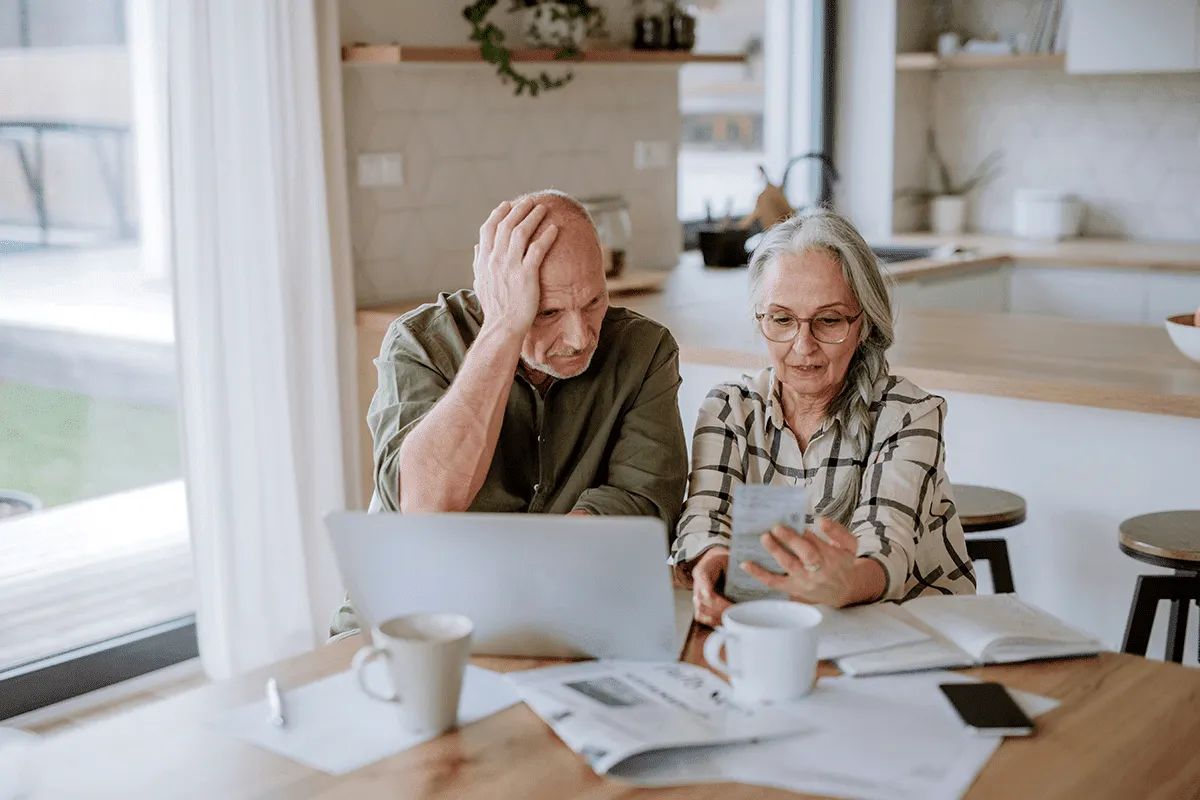 An older couple sits at a kitchen table looking concerned. The man holds his head while looking at a laptop, and the woman holds a sheet of paper—perhaps weighing their options for Retirement Living or considering MorningStar Senior Living Kirkland.
