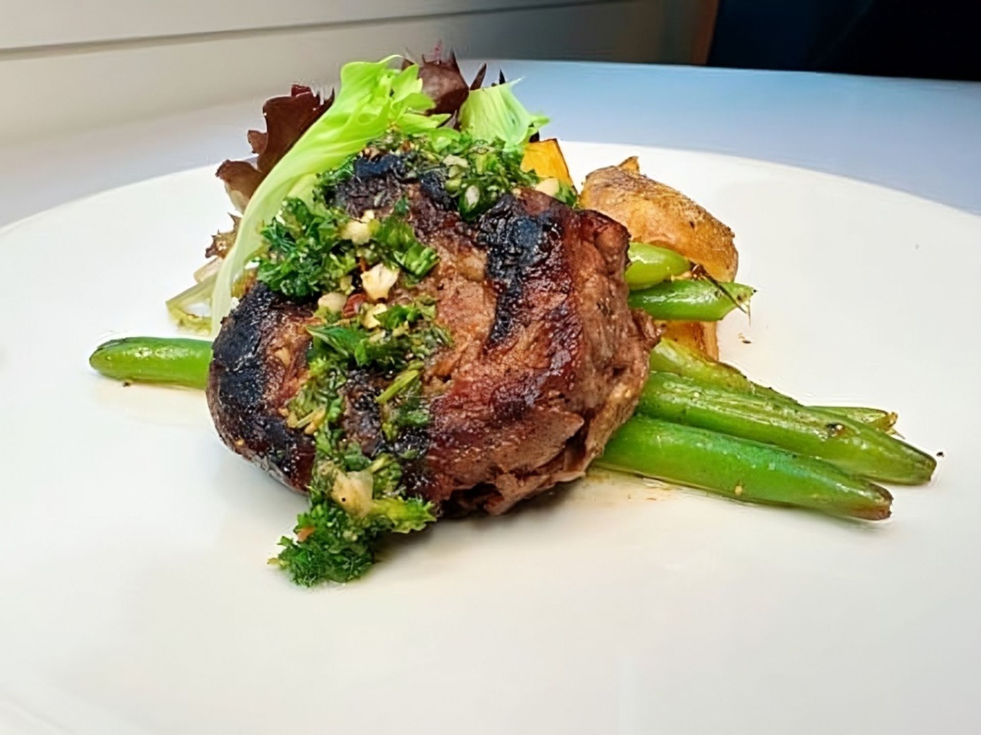 A grilled steak topped with chopped herbs and garlic is served on green beans, accompanied by roasted potato wedges and mixed salad leaves—a delicious offering at MorningStar Senior Living Kirkland perfect for Retirement Living.