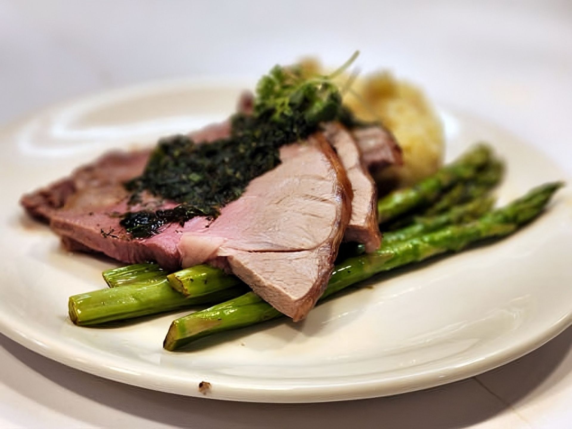Sliced roast lamb topped with green herb sauce served over asparagus spears, with a side of mashed potatoes—just one example of the delicious meals enjoyed at MorningStar Senior Living Kirkland for exceptional retirement living.