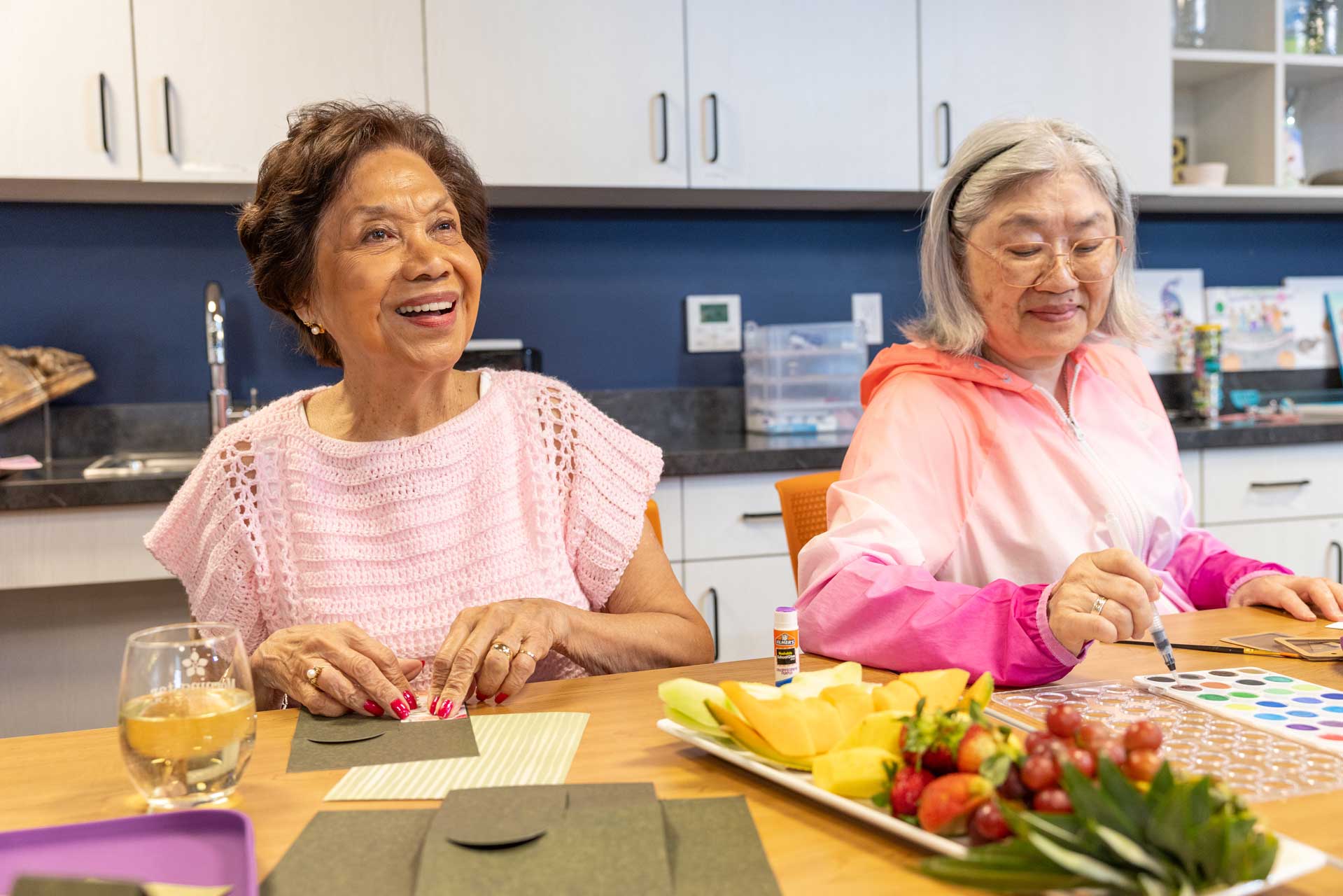 Two older women enjoy arts and crafts at a table in the bright, modern MorningStar Senior Living Kirkland community. One smiles while folding paper; the other draws with a marker beside a plate of fruit and cheese.