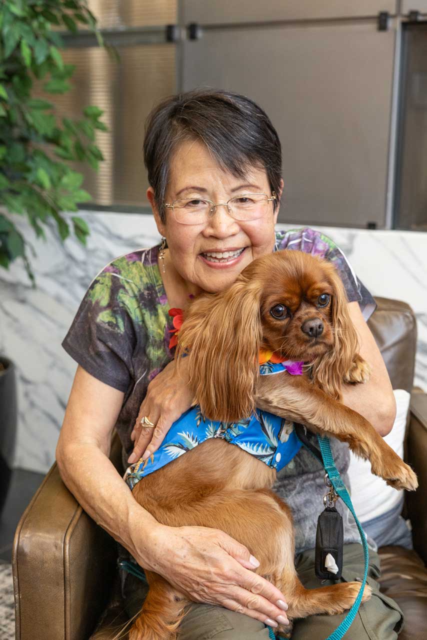 An older woman with glasses smiles while hugging a small brown dog in a blue harness. They are sitting on a brown chair indoors at MorningStar Senior Living Kirkland, with plants and marble decor creating a welcoming retirement living atmosphere.