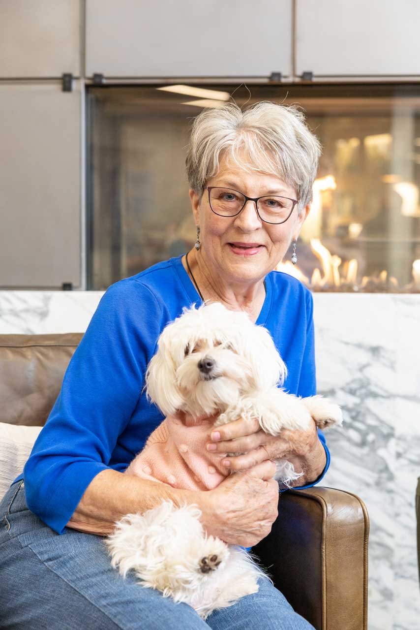 An older woman with gray hair and glasses, wearing a blue top, sits on a brown sofa and smiles while holding a small white dog in her lap. The cozy scene at MorningStar Senior Living Kirkland features a modern fireplace and marble wall in the background.