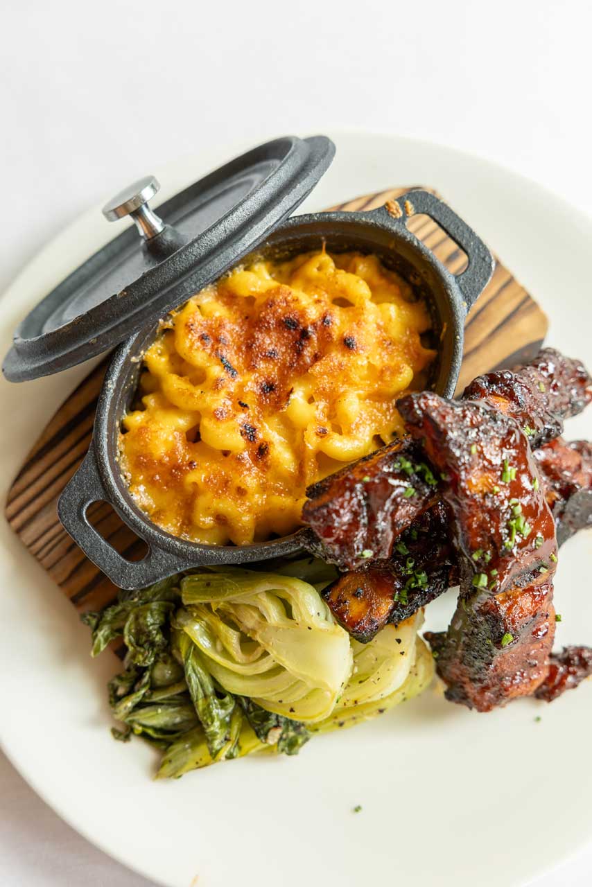 A plate with baked macaroni and cheese in a small cast iron pot, glazed barbecue ribs, and sautéed greens—all served on a white plate at MorningStar Senior Living Kirkland, where retirement living meets delicious dining.