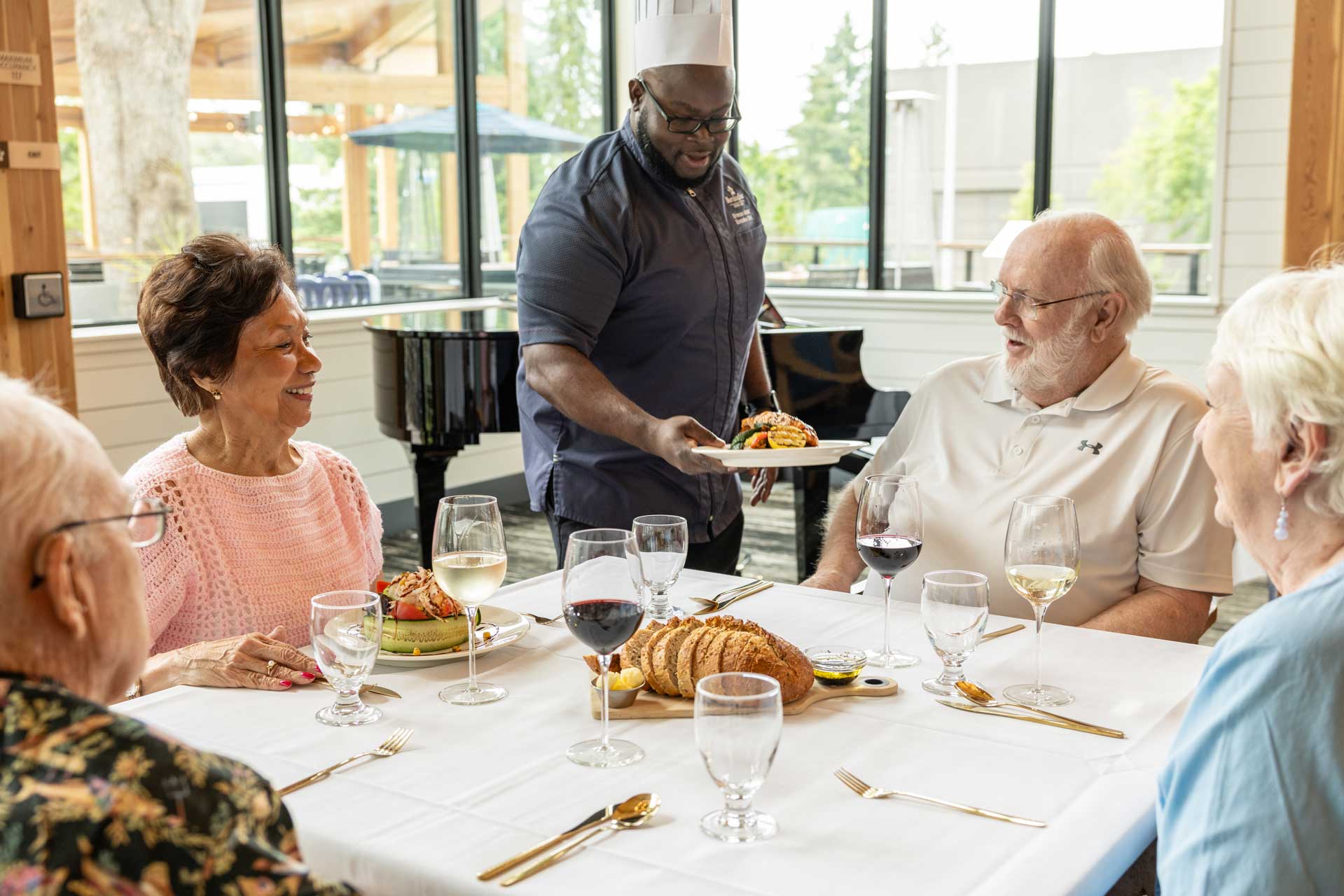A chef serves a plated meal to a smiling elderly man seated with three others in a bright, modern dining area at MorningStar Senior Living Kirkland, where retirement living means enjoying delicious food, wine, and bread together.