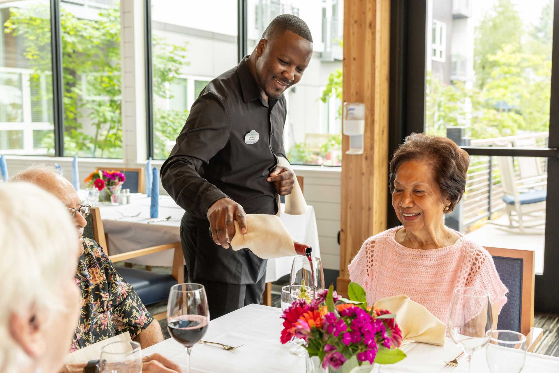 A waiter pours red wine for a smiling woman at a table set for dining with flowers and wine glasses, while two others sit across from her in the bright MorningStar Senior Living Kirkland restaurant.