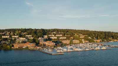 A marina filled with boats extends into a calm body of water, with buildings and houses nestled among trees on a hillside in the background—an inviting scene reminiscent of Retirement Living at MorningStar Senior Living Kirkland under a clear sky.
