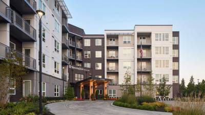 Modern multi-story apartment building with balconies, large windows, and an entrance canopy. An American flag is displayed above the entrance at MorningStar Senior Living Kirkland, with trees and landscaping along a curved driveway for retirement living.