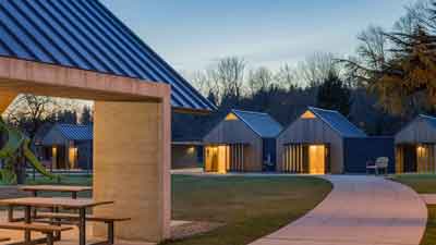 A park at dusk with a covered picnic area and several small, modern cabins in the background, all warmly lit from within—MorningStar Senior Living Kirkland offers inviting retirement living with curved sidewalks and distant trees.