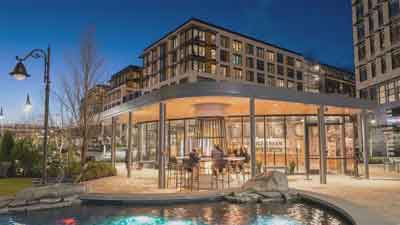 Modern building with large windows and outdoor seating is illuminated at dusk. In the foreground, a pool with rocks reflects colorful lights, while MorningStar Senior Living Kirkland offers residents vibrant retirement living surrounded by apartment buildings.