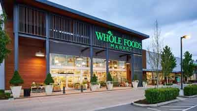 The exterior of a Whole Foods Market grocery store, featuring large windows, greenery, and the store's green logo above the entrance. Trees and potted plants line the front walkway near MorningStar Senior Living Kirkland Retirement Living.