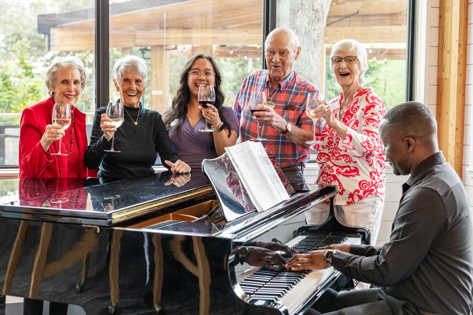 Five smiling older adults stand around a grand piano at MorningStar Senior Living Kirkland, holding wine glasses and raising them in a toast as a man plays the piano. Large windows in the background reveal an inviting retirement living scene.