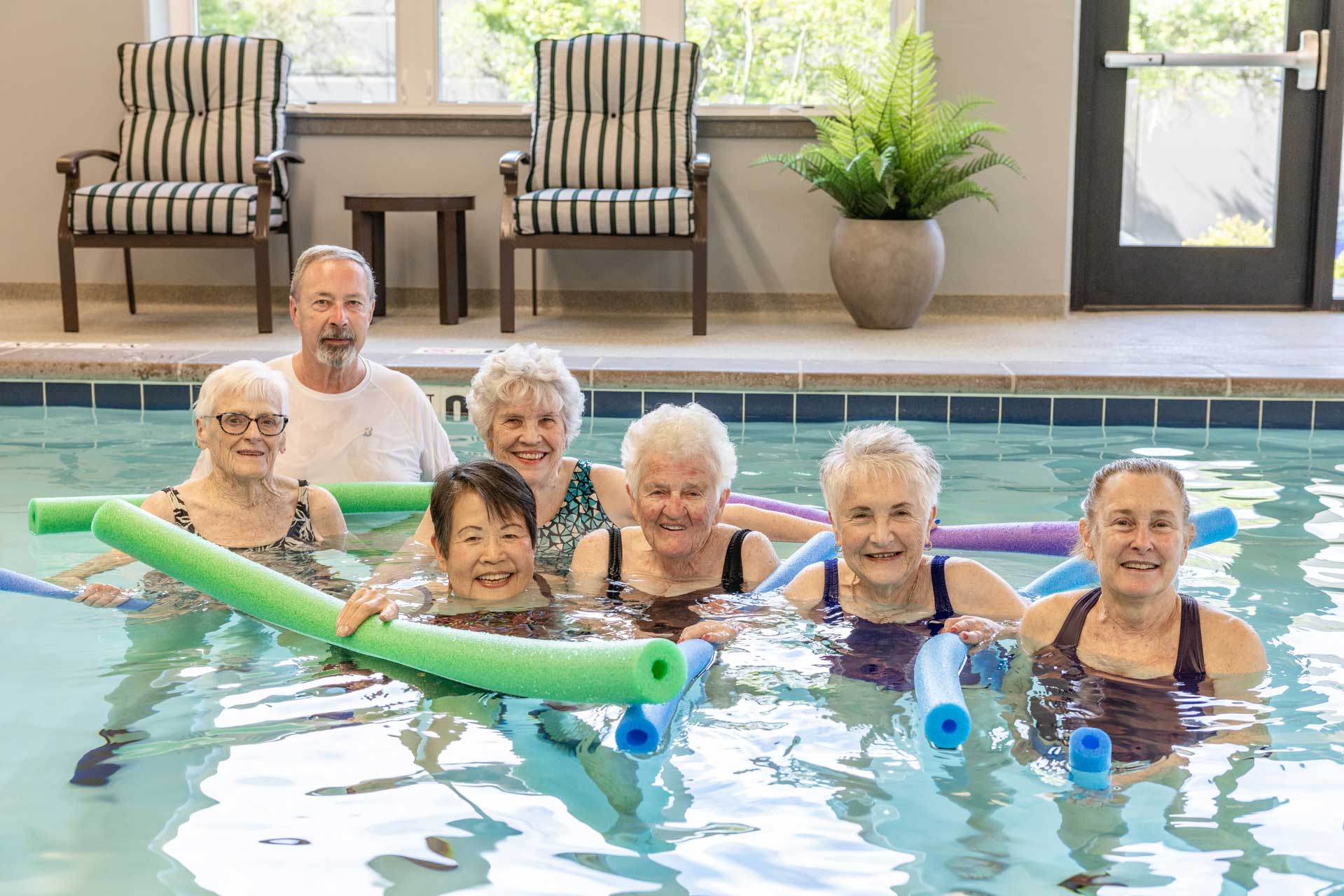 A group of seven smiling seniors, six women and one man, stand in an indoor pool at MorningStar Senior Living Kirkland, holding colorful pool noodles. Two striped chairs and a potted plant are in the background.