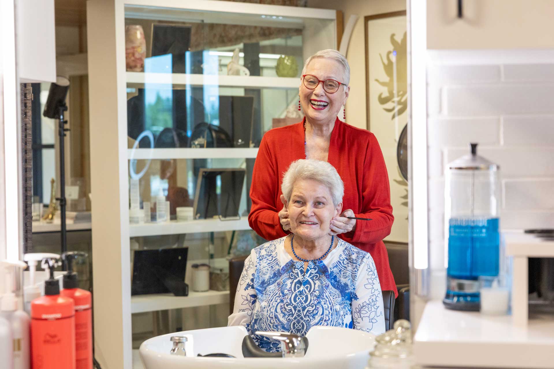 At MorningStar Senior Living Kirkland, two elderly women enjoy a brightly lit salon. One, standing and smiling in a red jacket, styles the hair of her friend, who sits in a white and blue top, beaming at the camera—retirement living at its finest.