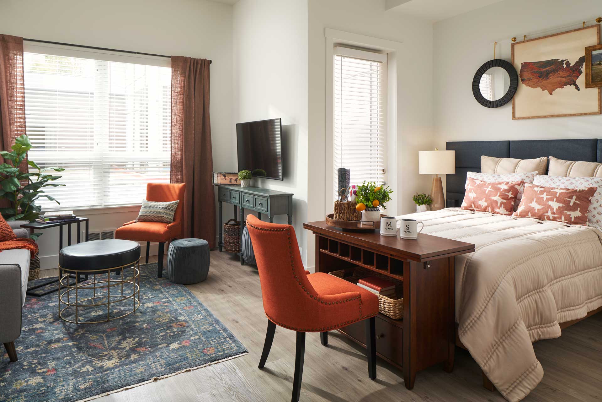 A cozy studio apartment at MorningStar Senior Living Kirkland features a bed, orange chair at a wooden desk, wall-mounted TV, sofa, large window with blinds, and decorative accents like a map, plants, and art on the walls—perfect for retirement living.
