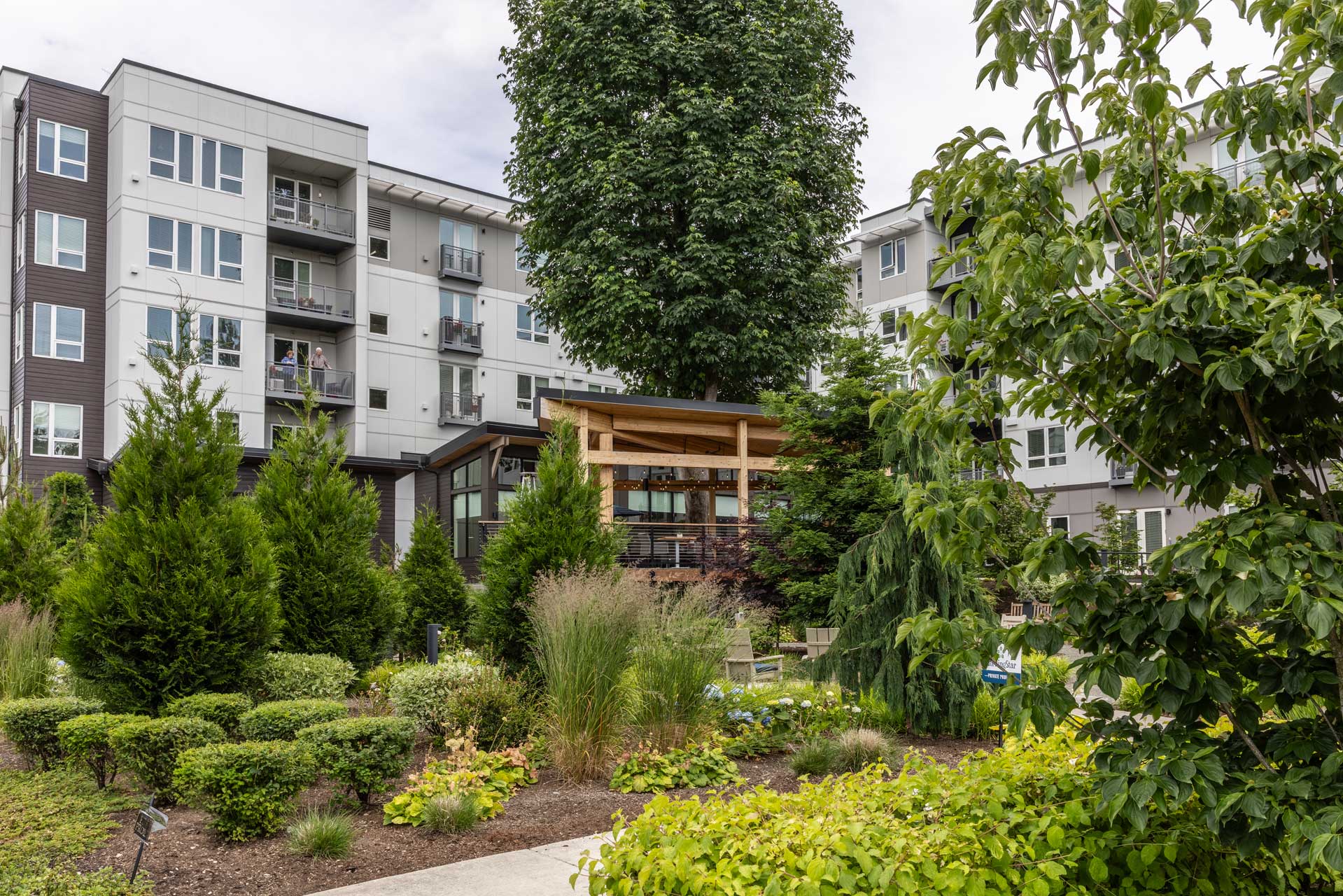 A modern apartment building with balconies at MorningStar Senior Living Kirkland overlooks a lush, landscaped garden featuring various shrubs, small trees, and a wooden pergola in the center—perfect for retirement living.