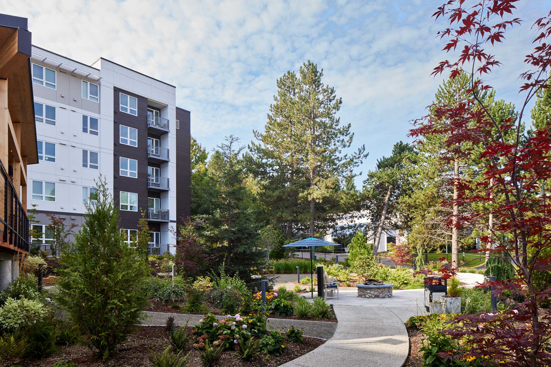 A landscaped courtyard with walking paths, colorful plants, trees, benches, and a fire pit offers inviting Retirement Living at MorningStar Senior Living Kirkland, surrounded by a modern multi-story apartment building under a partly cloudy sky.