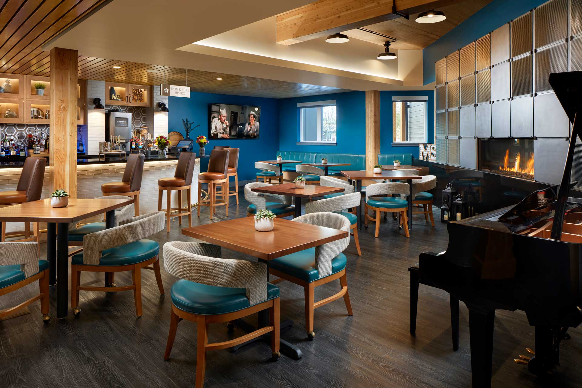 Cozy restaurant at MorningStar Senior Living Kirkland with wooden tables and blue cushioned chairs, a bar area with stools, modern fireplace, grand piano, and warm lighting—perfect for relaxing retirement living. Decor shelves and a TV add to the charm.