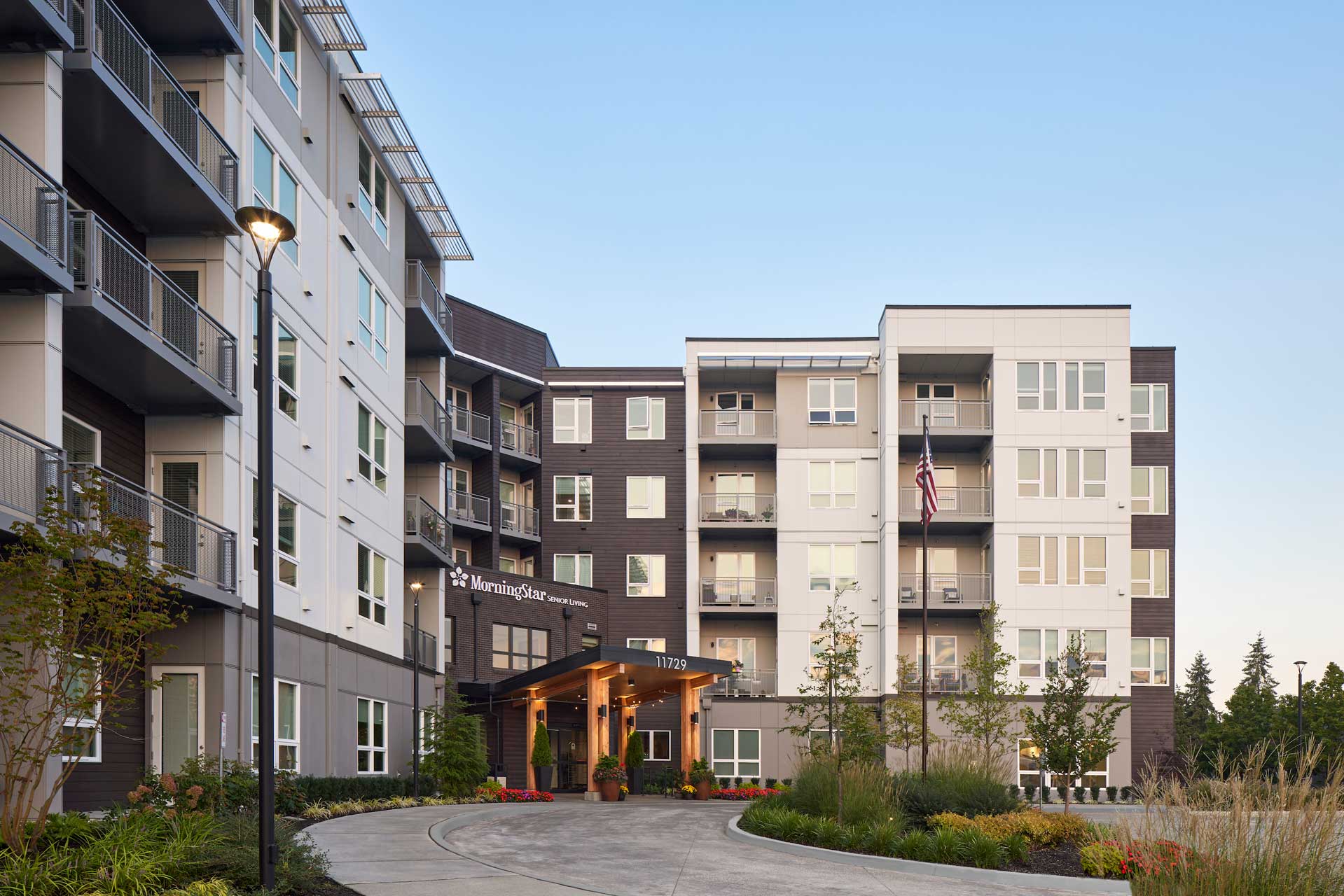 Modern multi-story apartment building with balconies, a covered entrance, and an American flag. MorningStar Senior Living Kirkland offers retirement living with a curved driveway, landscaped plants, and trees under a clear sky.
