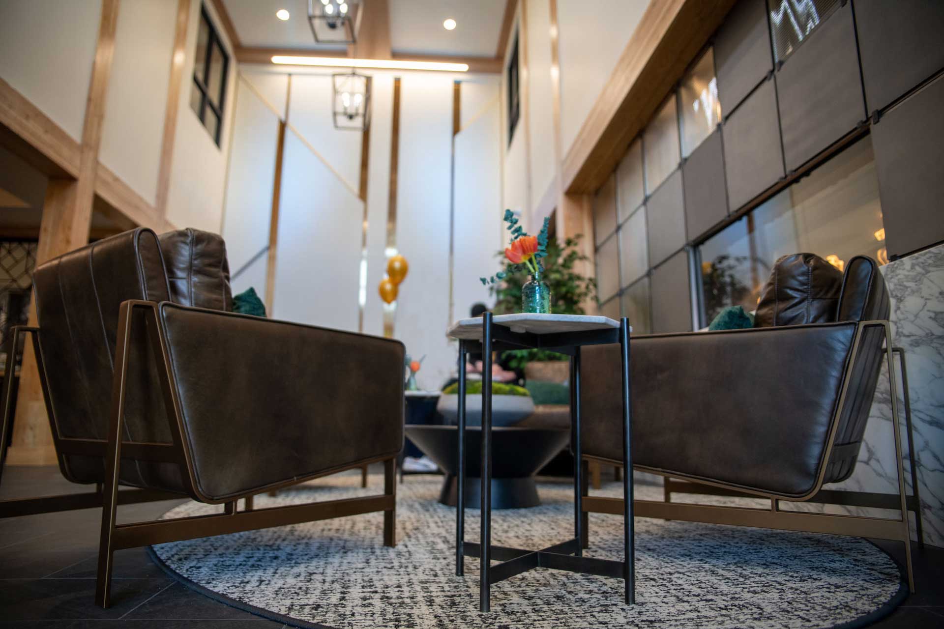 A modern lounge area at MorningStar Senior Living Kirkland features two leather armchairs, a small round table with a flower vase, and soft lighting in a spacious room with tall windows and wooden accents—perfect for comfortable retirement living.