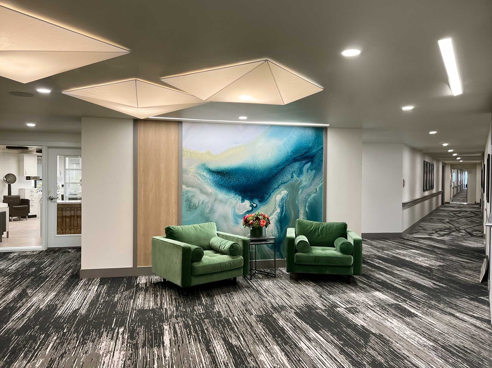 A modern hallway at MorningStar Senior Living Kirkland features patterned carpet, two green armchairs, a round table with a flower arrangement, large blue abstract wall art, geometric ceiling lights, and a visible corridor—perfect for elegant retirement living.
