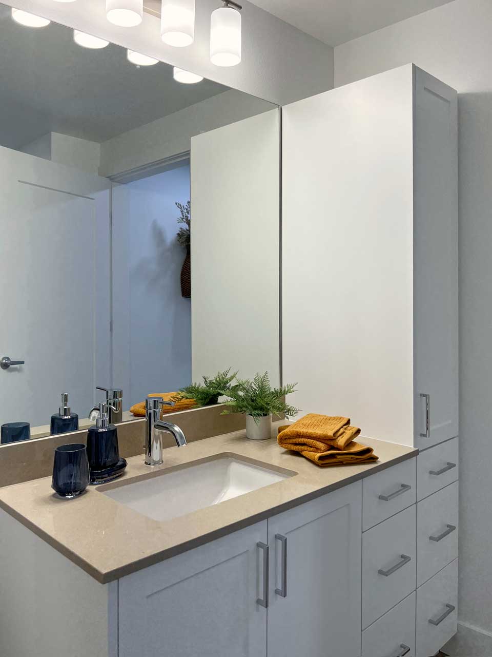 A modern bathroom at MorningStar Senior Living Kirkland features a beige countertop, white cabinets, a sink with silver fixtures, a mirror, dark soap dispensers, a small potted plant, and neatly folded mustard yellow towels—perfect for retirement living.