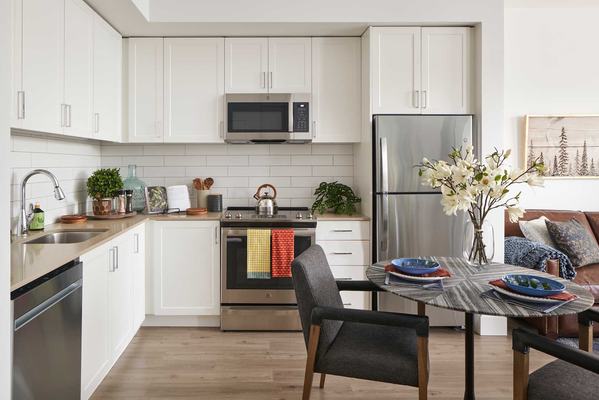 Modern kitchen with white cabinets, stainless steel appliances, and a round dining table set for two at MorningStar Senior Living Kirkland. The living area with a sofa is partly visible in this inviting Retirement Living space.