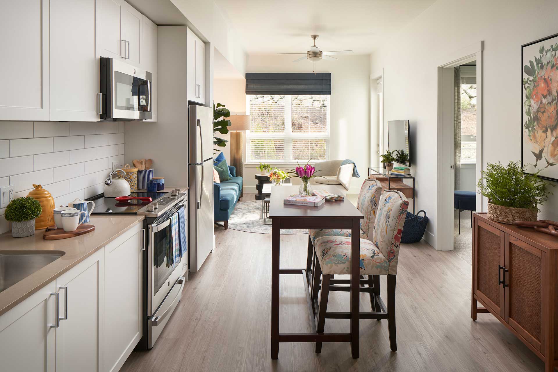 Modern apartment kitchen with white cabinets and stainless steel appliances, perfect for Retirement Living at MorningStar Senior Living Kirkland. Leads to a bright living room with blue sofa, round table, large window, and two floral chairs by the high dining table.