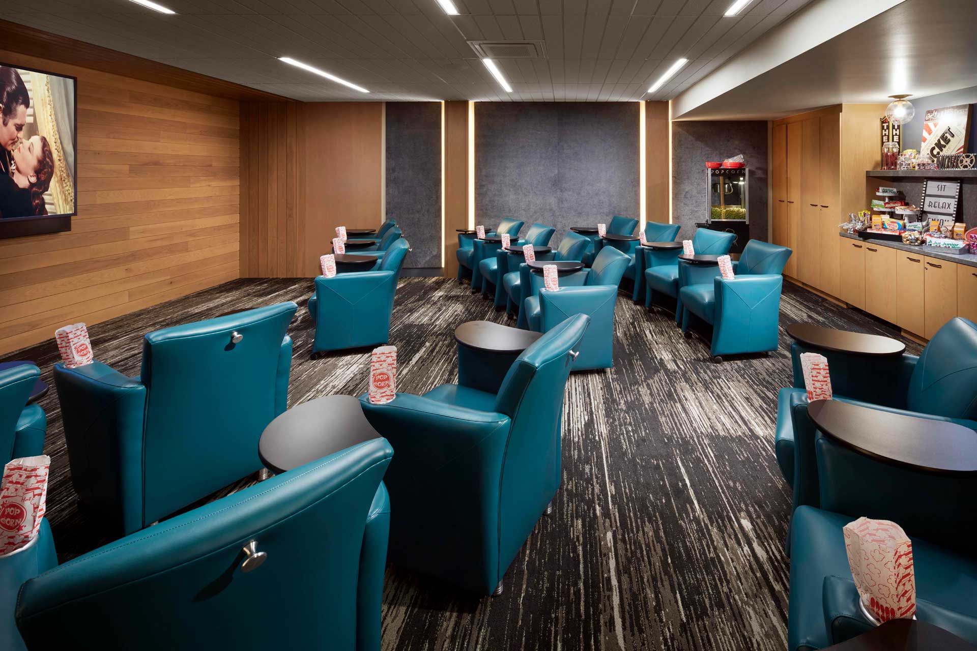 A modern private movie screening room at MorningStar Senior Living Kirkland features teal armchairs in rows, each with a tub of popcorn. A large screen shows a film, while snacks line the back wall—perfect for retirement living comfort and entertainment.