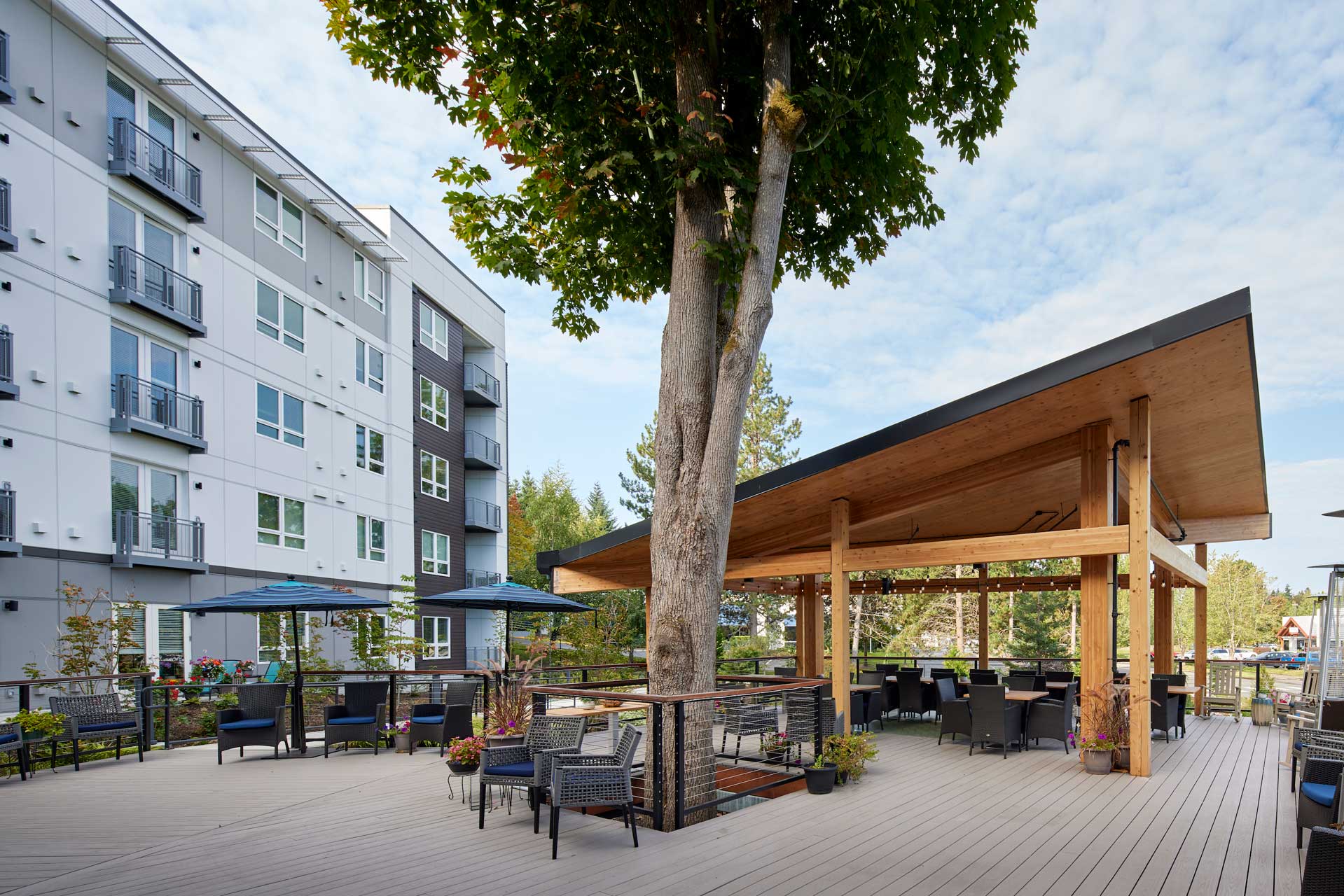 Outdoor patio with modern seating, shaded tables, and a large tree growing through a wooden pavilion. Apartment building in the background—all part of MorningStar Senior Living Kirkland—lush greenery and open blue sky complete this Retirement Living oasis.