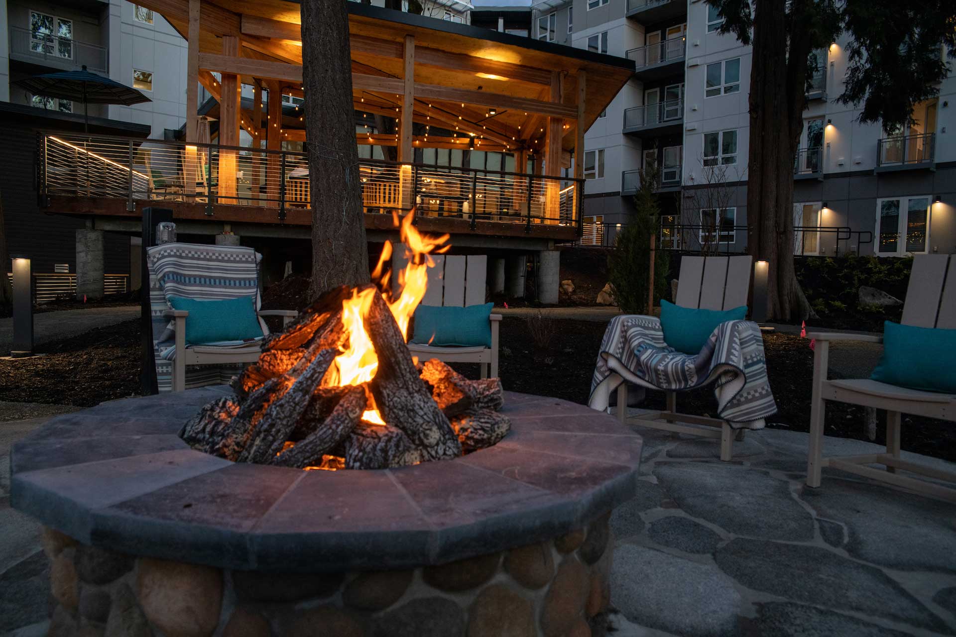 A cozy outdoor fire pit with burning logs, surrounded by cushioned chairs and blankets, sits in front of a warmly lit pavilion at MorningStar Senior Living Kirkland, offering inviting retirement living near modern apartment buildings in the evening.