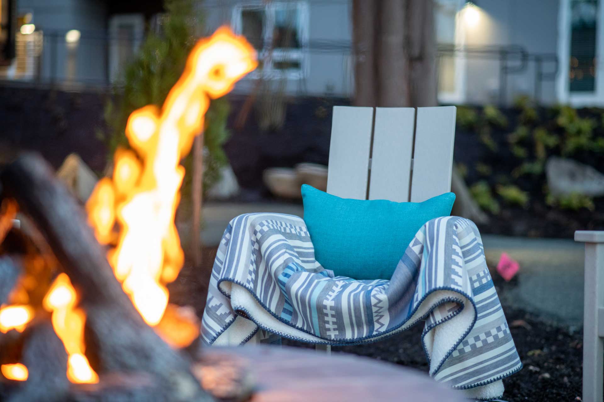 A wooden chair with a turquoise pillow and patterned blanket sits near a campfire outdoors at MorningStar Senior Living Kirkland, with a building and greenery blurred in the background, creating a cozy retirement living scene.