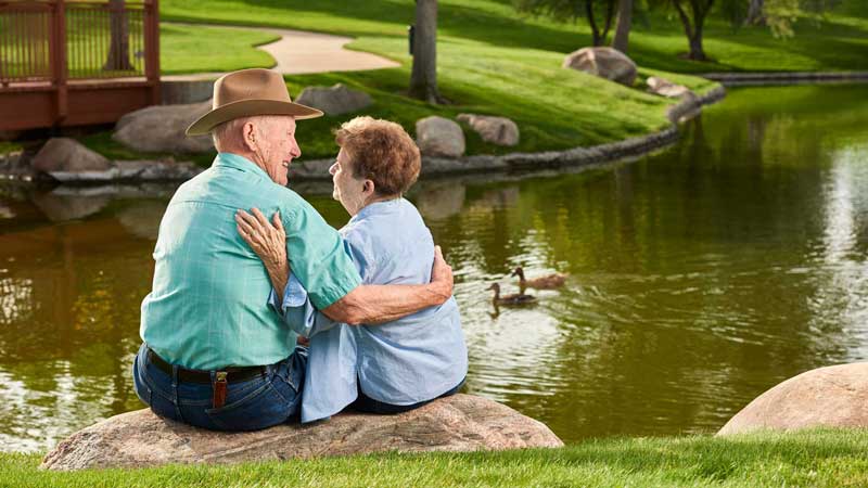 An elderly couple sits on a rock by a pond at MorningStar Senior Living Kirkland, embracing and smiling as ducks swim nearby and a grassy park forms the tranquil backdrop—a perfect moment of retirement living.