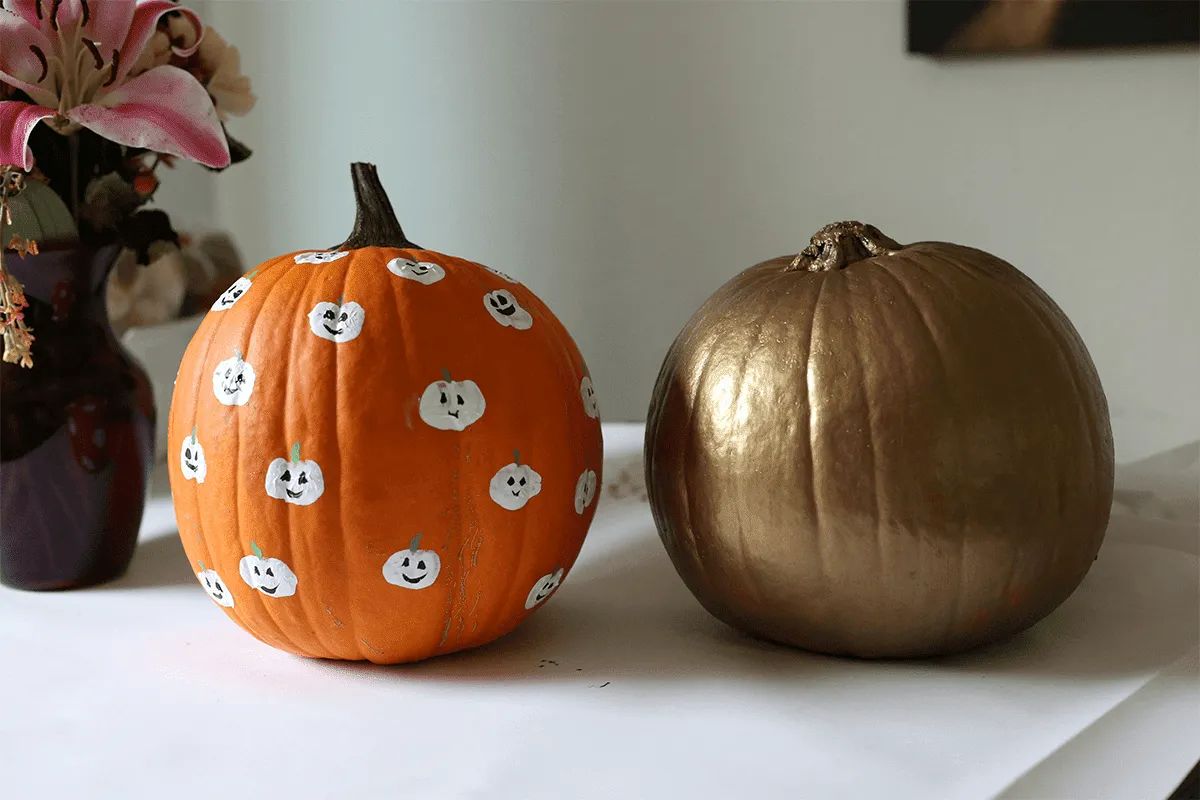 Two pumpkins on a table at MorningStar Senior Living Kirkland: one orange pumpkin with small white smiling faces and one painted gold. In the background on the left, pink flowers in a vase add cheer to this Retirement Living scene.