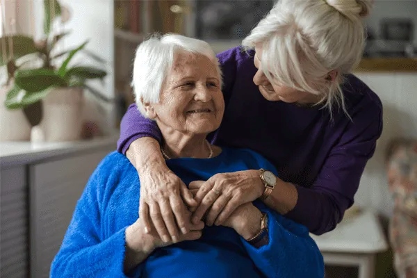 An elderly woman smiles while sitting, as a younger woman with white hair hugs her from behind, both sharing a warm, affectionate moment indoors at MorningStar Senior Living Kirkland.