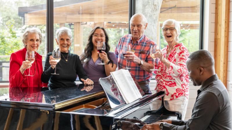Five smiling older adults stand around a grand piano at MorningStar Senior Living Kirkland, holding wine glasses and raising them in a toast as a man plays the piano. Large windows in the background reveal an inviting retirement living scene.