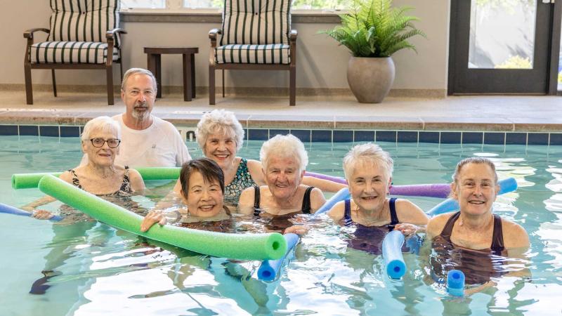 A group of six older women and one older man smile while standing in a pool at MorningStar Senior Living Hillsboro, holding colorful pool noodles, with chairs, a plant, and windows visible in the background—enjoying vibrant retirement living.