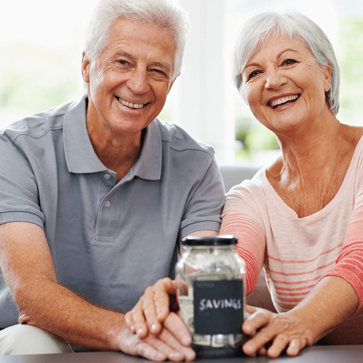 Smiling older couple sitting together, holding a glass jar labeled "SAVINGS" filled with cash, suggesting financial planning or saving for the future at MorningStar Senior Living Kirkland.