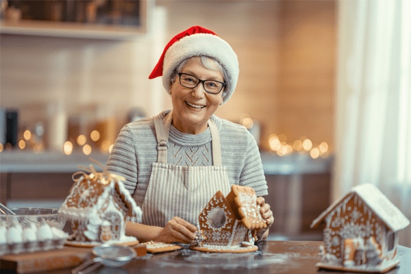 An elderly woman wearing a Santa hat and apron smiles while decorating a gingerbread house in a festive kitchen with holiday lights in the background, enjoying the warmth and joy of Retirement Living at MorningStar Senior Living Kirkland.