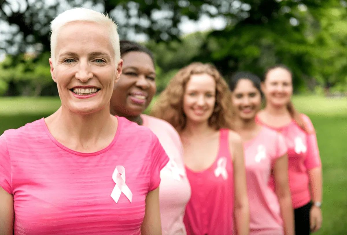Five women from MorningStar Senior Living Kirkland wear pink tops with ribbon pins, standing outdoors in a line and smiling. The woman in front has short blonde hair. The green, leafy background hints at a vibrant retirement living community.