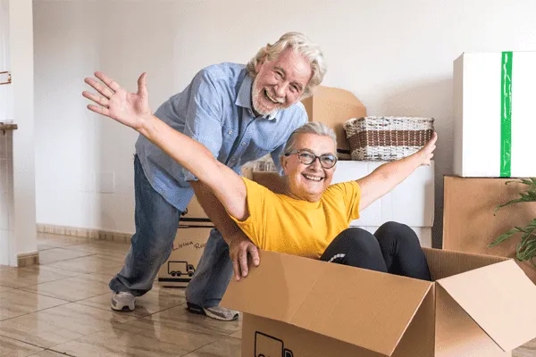 An older man smiles and stands behind an older woman who sits playfully in a cardboard box, arms outstretched. Surrounded by moving boxes indoors, they celebrate a joyful new chapter at MorningStar Senior Living Kirkland.