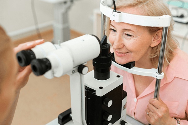 A woman sits at an eye examination device, smiling, while an optometrist prepares to check her vision in the MorningStar Senior Living Kirkland retirement living clinic.