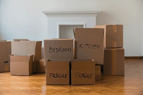 Several cardboard moving boxes labeled “BEDROOM,” “KITCHEN,” and “FRAGILE” are stacked on a wooden floor in front of a white wall and fireplace, suggesting a home move to MorningStar Senior Living Kirkland or a new retirement living space.