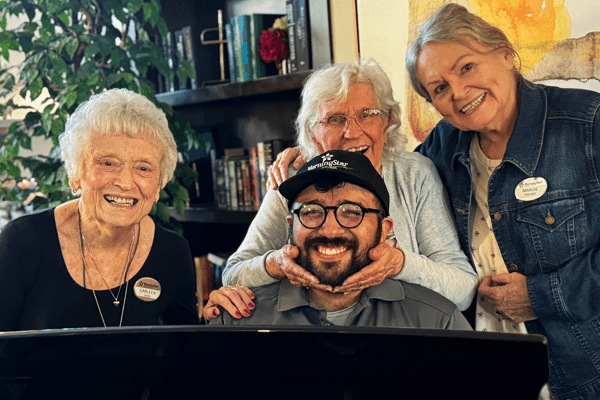 Three smiling elderly women stand behind a seated man wearing glasses and a cap. Their joy shines in this cozy indoor setting at MorningStar Senior Living Kirkland, with plants and books reflecting vibrant retirement living.