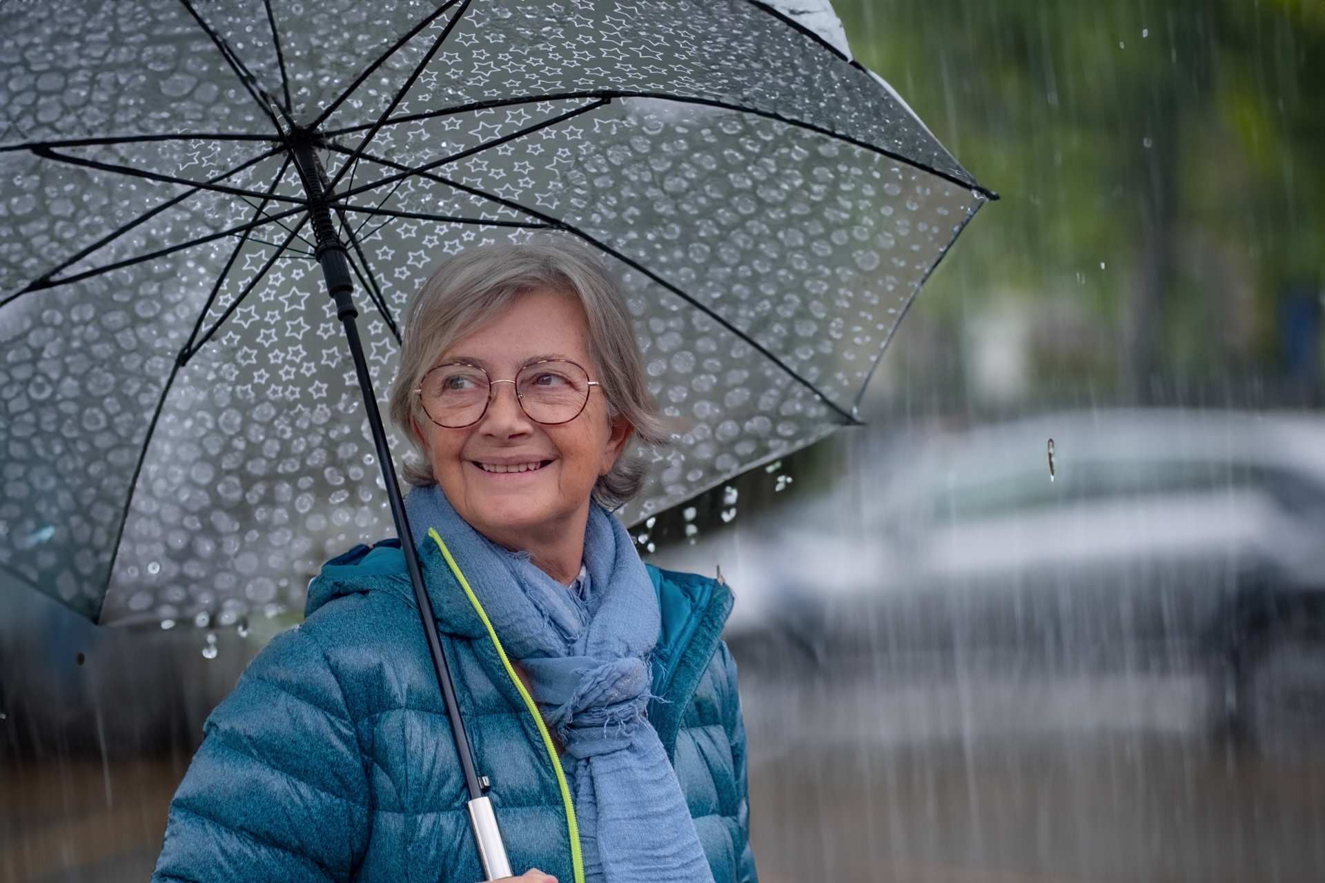 A smiling senior woman standing under an umbrella in the rain.