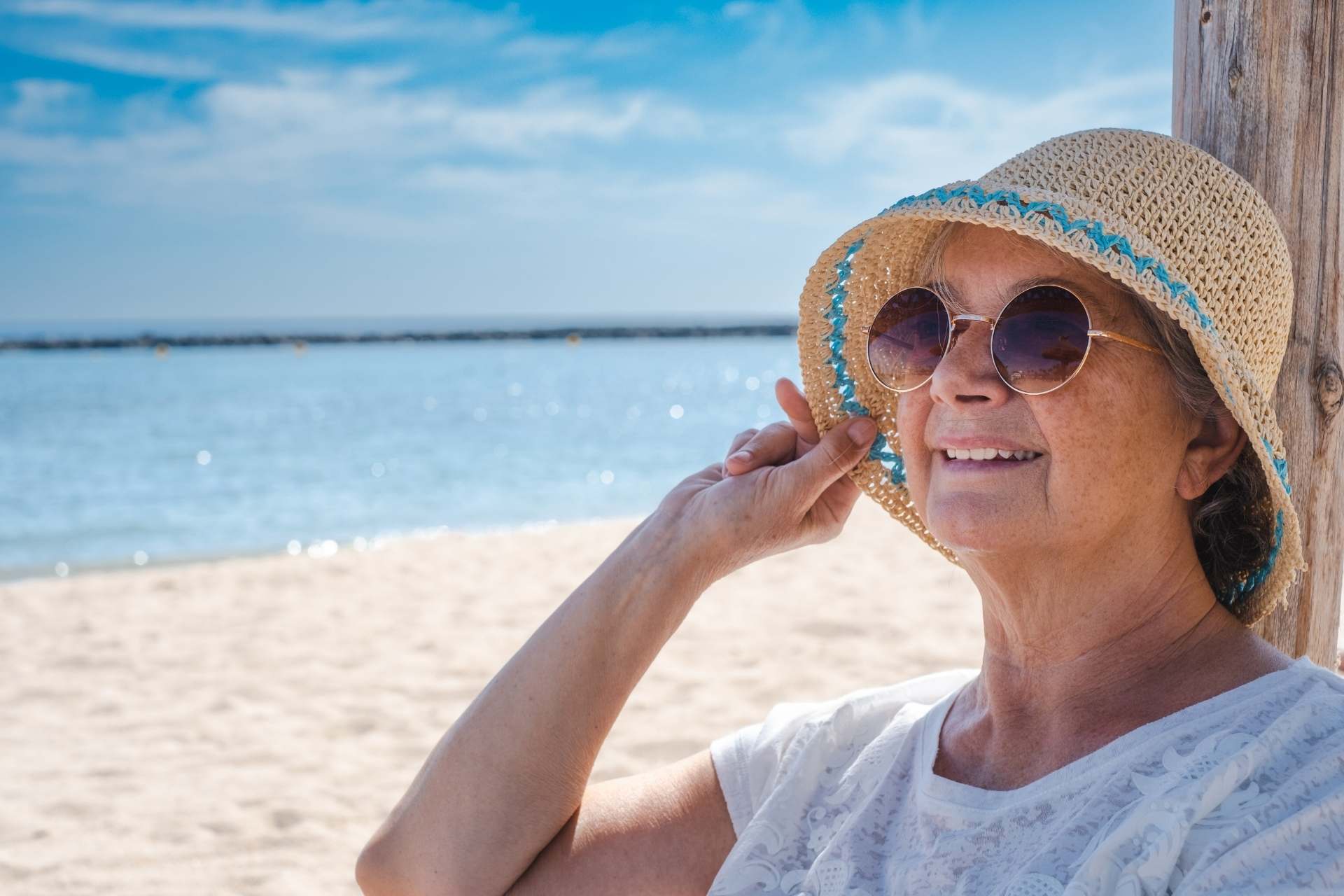 smiling-senior-woman-with-summer-hat-and-sunglasses Smiling senior woman enjoying a beach day in the shade.